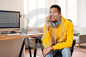 Cheerful black teen guy with headphones on neck talking on phone at home