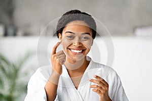 Cheerful black woman applying moisturizing cream on her cheek