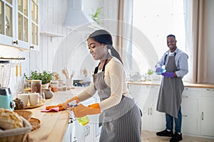 Cheerful black woman cleaning kitchen table, her boyfriend washing dishes on background, blank space