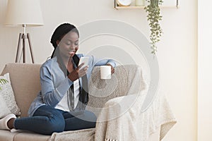 Cheerful black woman using smartphone and drinking coffee on couch