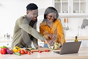 Cheerful Black Young Couple Using Laptop While Cooking Healthy Food In Kitchen