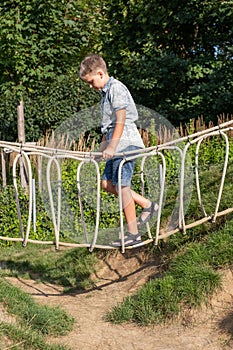 Cheerful blond boy walks on rope bridge on a wooden playground in a public park.