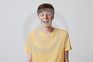 Cheerful blond young man with short haircut and metal braces on teeth wears yellow t shirt and looks happy  over white