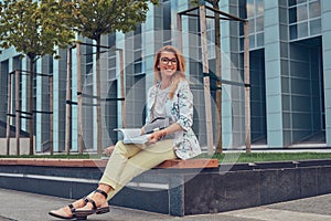 Charming blonde female in modern clothes, studying with a book, sitting on a bench in the park against a skyscraper.