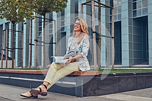 Charming blonde female in modern clothes, studying with a book, sitting on a bench in the park against a skyscraper.