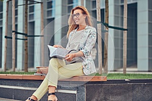 Charming blonde female in modern clothes, studying with a book, sitting on a bench in the park against a skyscraper.