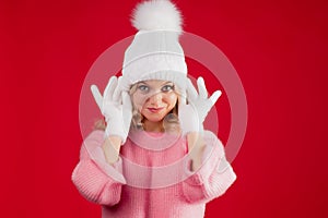 A cheerful blonde in a warm pink sweater and a white hat with gloves on a red background.