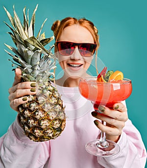 Cheerful blonde woman in modern red sunglasses handed us demonstrates a strawberry margarita cocktail and a big pineapple