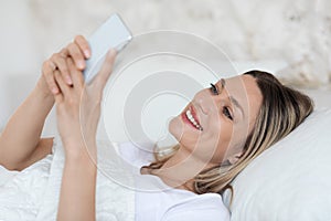 Cheerful blonde woman resting in bed with cell phone