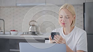 Cheerful blonde woman says bye on phone call when sitting at laptop in kitchen