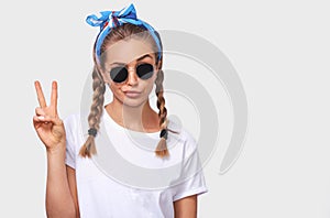 Cheerful blonde young woman wearing trendy sunglasses, white t-shirt and blue headband, making a duck face and showing peace sign
