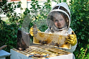 Cheerful boy beekeeper in protective suit near beehive. Honeycomb with honey. Organic food concept. The most useful photo