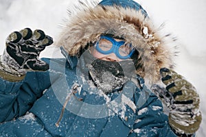 Cheerful boy in blue glasses is lying on the snow and looking at the camera