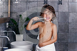 Cheerful boy with brown curly hair in white towel cleans his teeth using toothpaste and toothbrush in the bathroom.