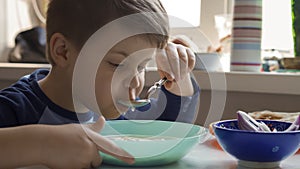 Cheerful boy eats soup with a spoon while sitting at the kitchen table. Hungry preschooler dines at home