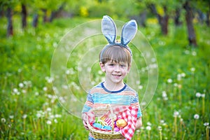 Cheerful boy holding basket full of colorful easter eggs standing on the grass in the park after egg hunt