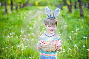 cheerful boy holding basket full of colorful easter eggs standing on the grass in the park after egg hunt