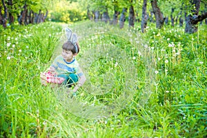 cheerful boy holding basket full of colorful easter eggs standing on the grass in the park after egg hunt