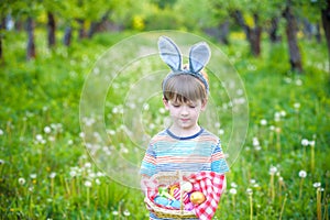 cheerful boy holding basket full of colorful easter eggs standing on the grass in the park after egg hunt