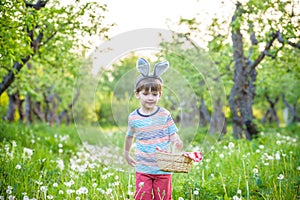 Cheerful boy holding basket full of colorful easter eggs standing on the grass in the park after egg hunt