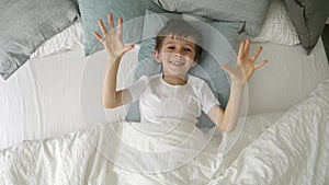 A cheerful boy lying in bed and opening his blanket with a smile directed at the camera, showing joy and a playful spirit.