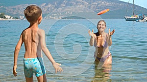 Cheerful boy with mother throwing and catching frisbee disk while relaxing in the calm sea water at beach. Family