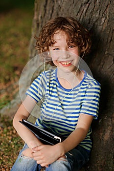 The cheerful boy sits under a tree with tablet on lap and puts out the tongue.