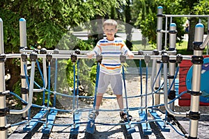 Cheerful boy walks on a rope bridge on a playground in the park.