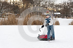 Cheerful boy in winter jumpsuit and bright hat runs through snow with tubing. Child on winter vacation is having fun