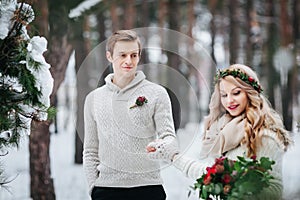 Cheerful bride and groom in beige knitted pullovers are walking in snowy forest. Selective focus on the groom. Artwork photo