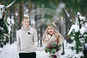 Cheerful bride and groom in beige knitted pullovers are walking in snowy forest. Selective focus on bride. Artwork