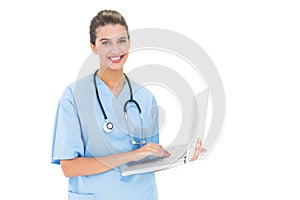 Cheerful brown haired nurse in blue scrubs using a laptop