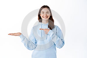 Cheerful brunette girl pointing at her palm holding copyspace, showing item on display in her hand, standing against