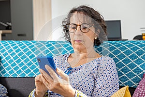 Cheerful brunette senior woman using smartphone while sitting on sofa