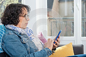 Cheerful brunette senior woman using smartphone while sitting on sofa