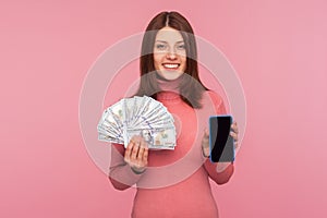 Cheerful brunette woman in pink sweater holding dollars banknotes and cell phone with mock up blank display, online payments,