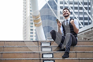 Cheerful Business Man Celebrating Success with mobile phone sitting on the stairs in urban city outdoors, looking up , good news,