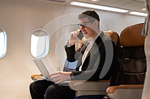 Cheerful businessman in airliner using laptop computer while talking on cellphone