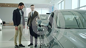 Cheerful businessman buying car in showroom talking to sales agent looking at auto. Focus on car mirror
