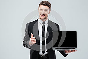 Cheerful businessman holding blank screen laptop and showing thumbs up