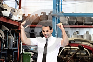 Cheerful businessman raising hands up to celebrate success at auto spare parts store, surrounded by numerous secondhand engine