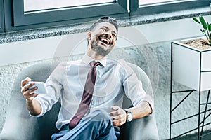 cheerful businessman sitting in armchair and laughing during video chat on smartphone.