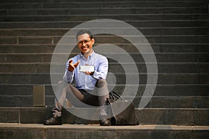 Cheerful businessman sitting on stairs of office building and showing smartphone with blank screen