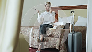 Cheerful businesswoman talking mobile phone and writing notes in notepad while sits on bed in hotel room. Travel