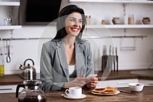 Cheerful businesswoman using smartphone near breakfast