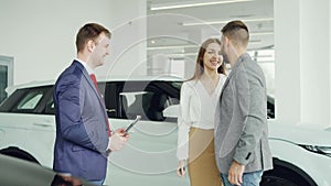 Cheerful car dealership salesman is shaking hands with young buyer handsome man then giving key fob to his happy wife