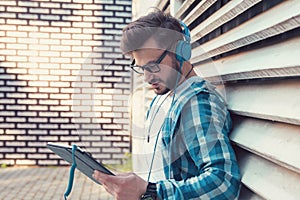 Cheerful casual man in denim shirt and headphones standing outdoors and surfing tablet happily