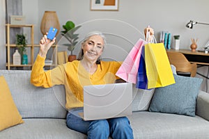 Cheerful caucasian elderly lady sit on sofa with laptop, shows a lot of packages with purchases and credit card