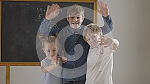 Cheerful Caucasian friends or siblings waving looking at camera standing in classroom at chalkboard. Positive happy boys
