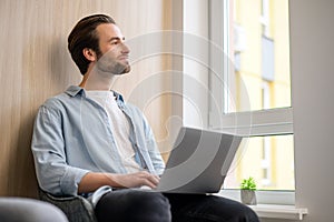 Cheerful caucasian guy sits on sofa in modern living room alone and using trendy laptop for net searching, web surfing